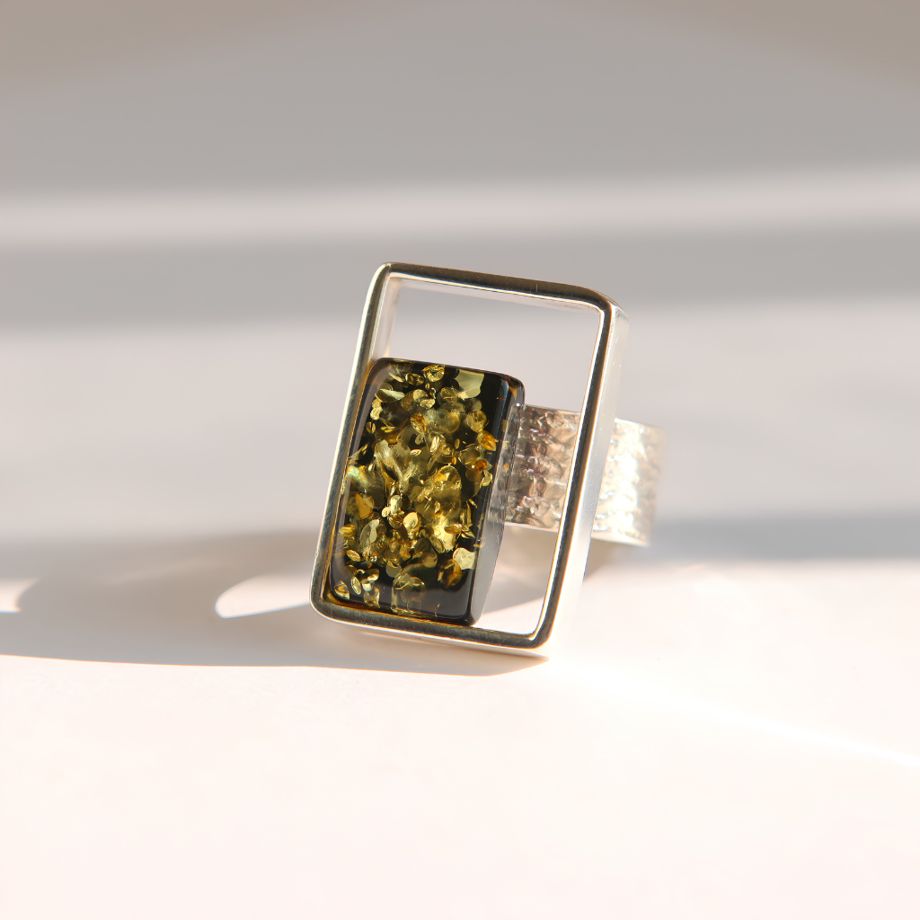 A silver amber ring with a rectangular frame and a central green amber inlay containing gold and black flecks, photographed on a light surface with soft shadows.