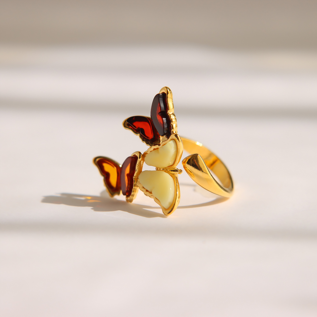 A gold amber butterfly ring with cream-colored amber wings and red accents, photographed on a warm, light beige background with soft shadows.