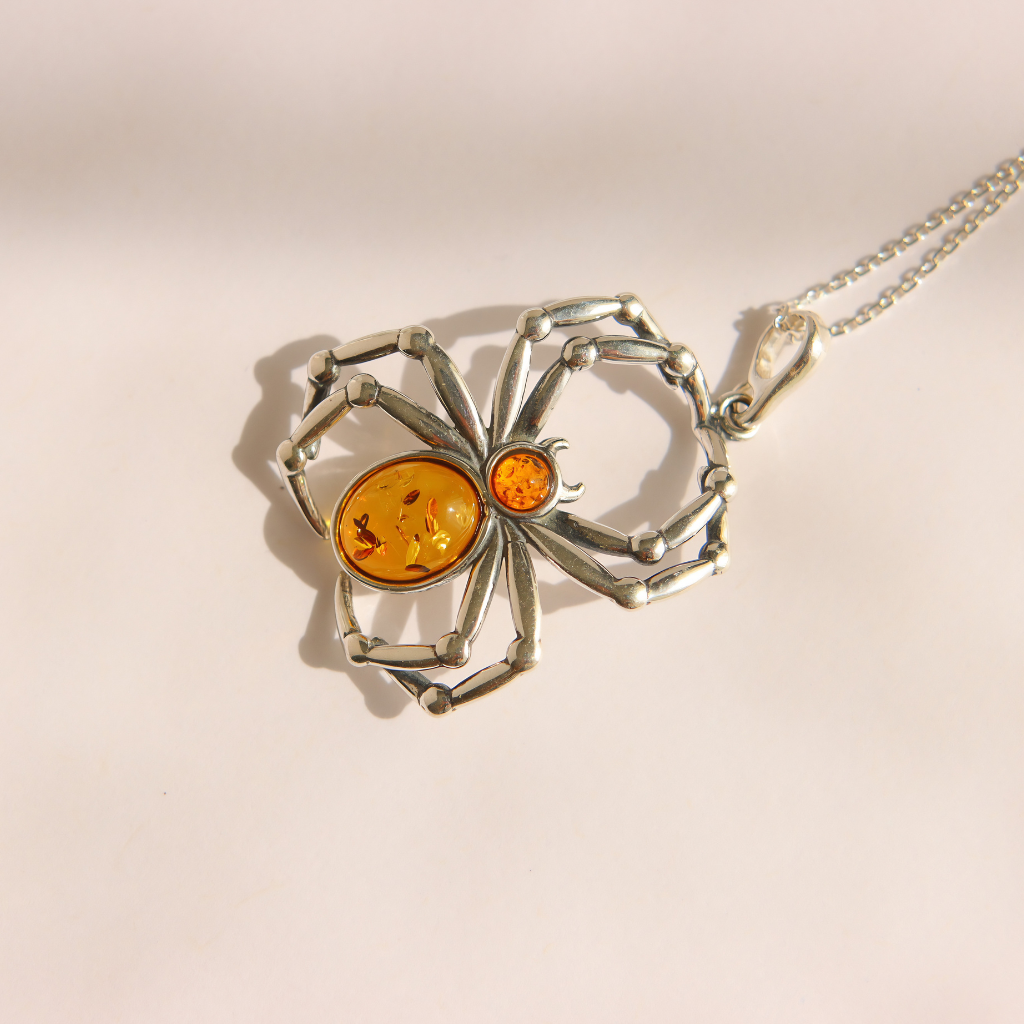A amber spider-shaped pendant necklace made of silver-toned metal with Baltic amber details, shown on a matching silver chain against a plain light background.