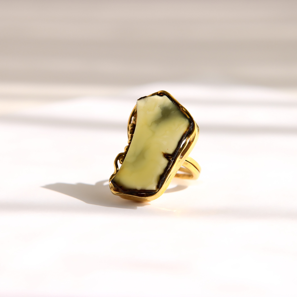 A gold amber statement ring featuring a large, irregularly shaped pale green stone with dark natural veining, photographed on a clean white surface with soft shadows.