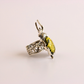 A silver green amber ring featuring a large, polished yellow-green beetle-shaped gemstone, photographed on a clean, light pink background.