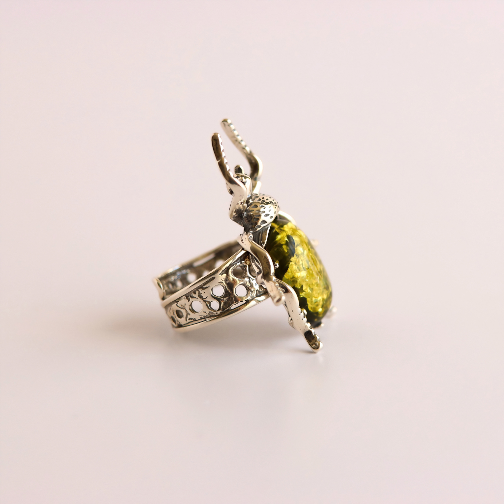 A silver green amber ring featuring a large, polished yellow-green beetle-shaped gemstone, photographed on a clean, light pink background.