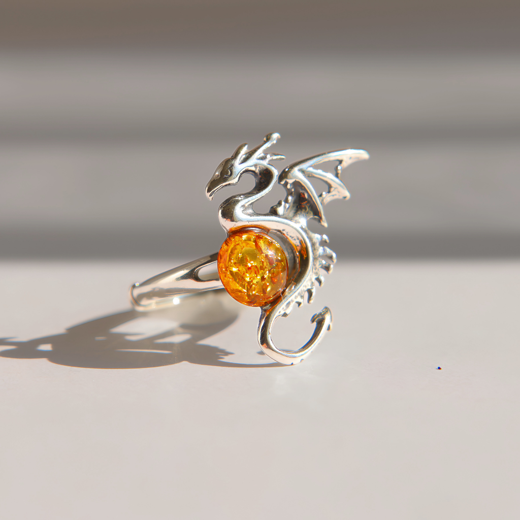 A silver amber dragon ring with a round, glittering amber-orange gemstone, photographed on a light gray surface with soft shadows.