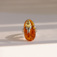 An oval amber ring featuring a small silver-toned metal figure inside, set against a warm off-white background with soft shadows.