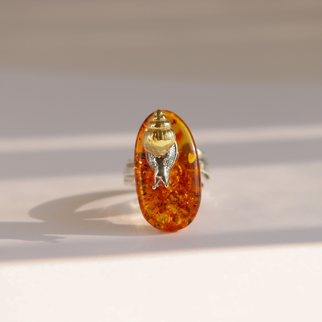 An oval amber ring featuring a small silver-toned metal figure inside, set against a warm off-white background with soft shadows.
