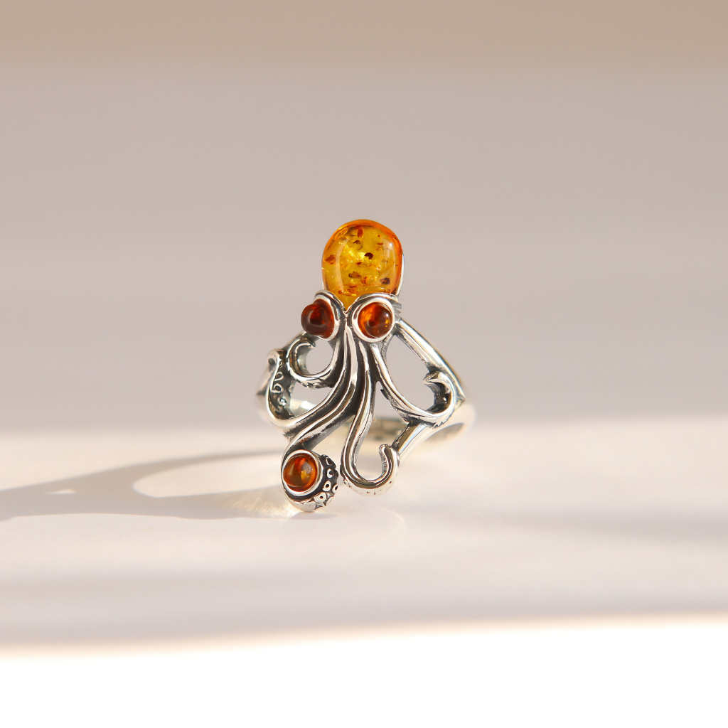 An ornate silver amber octopus ring featuring a large oval amber top stone and three smaller round amber accent stones, photographed on a clean, light beige background.