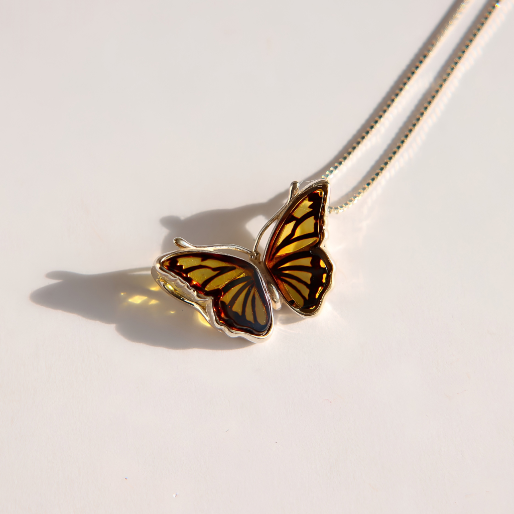 A silver amber butterfly pendant necklace with yellow and orange wings featuring black veins, resting on a white surface with a soft shadow.