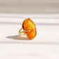 A gold amber ring featuring a large, organic-shaped orange amber cabochon gemstone, photographed on a clean white surface with soft, diagonal shadows.
