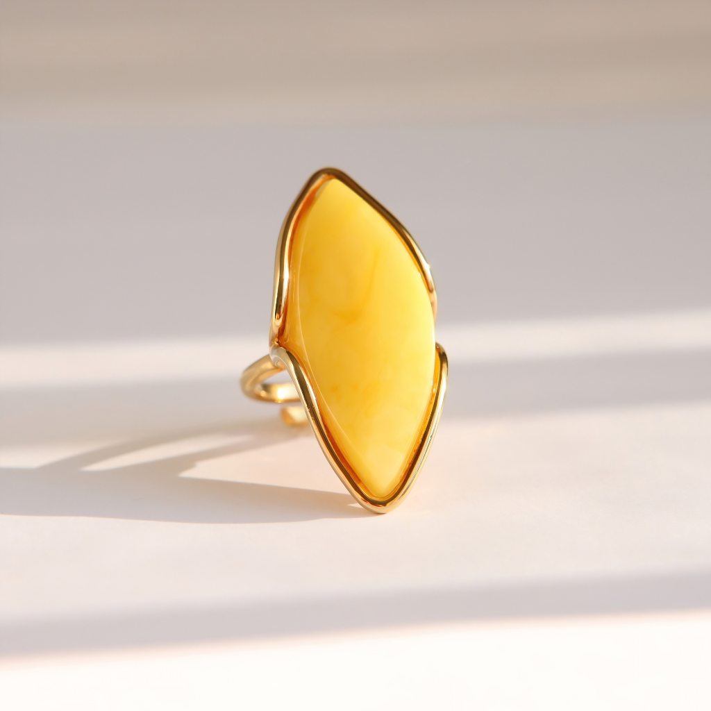 A gold butterscotch amber ring featuring a large, organic-shaped yellow stone, photographed on a light beige surface with soft shadows.