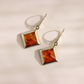A pair of silver drop amber earrings with square amber-orange gemstone pendants, displayed on a light beige surface with soft shadows.
