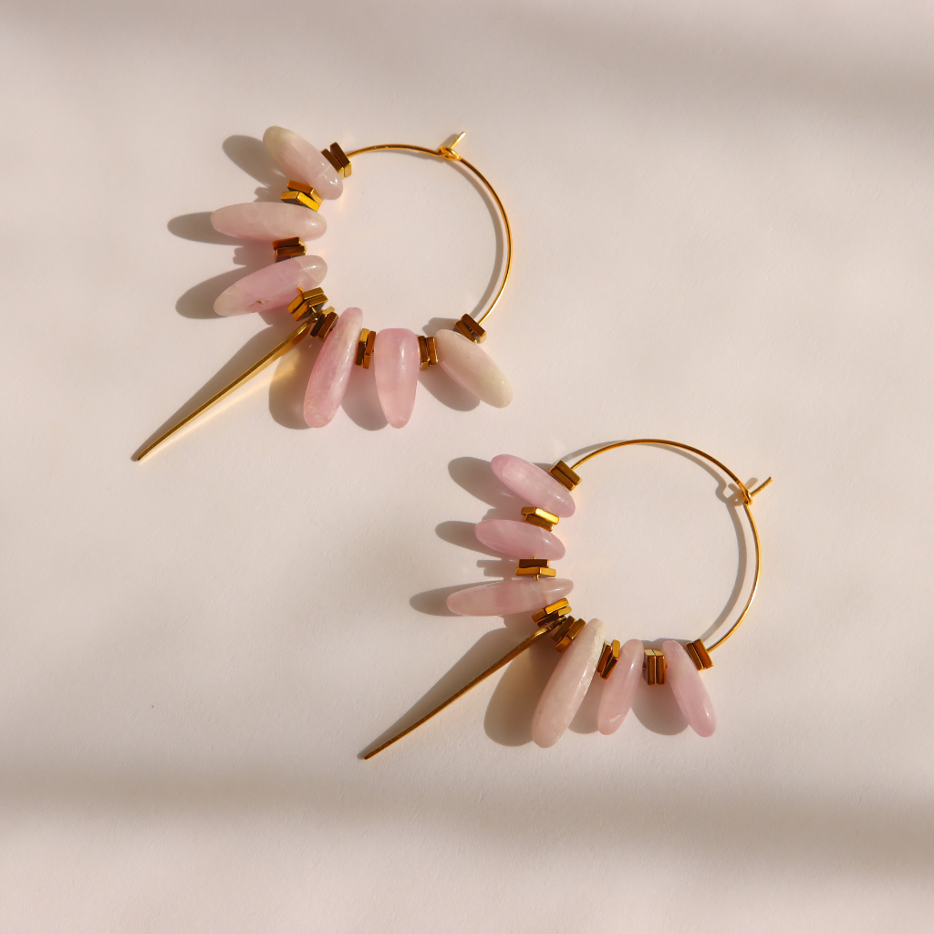 A pair of gold kunzite dagger hoop earrings with dangling pink stone beads, photographed on a warm off-white background with soft shadows.
