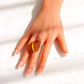 A hand with a French manicure wearing a large marquise-cut amber gemstone statement ring with a gold band, set against a bright white background with soft shadows.