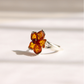 A silver amber flower ring featuring a flower-shaped setting with amber-colored stones and a small dark red center stone, photographed on a light beige surface with soft shadows.