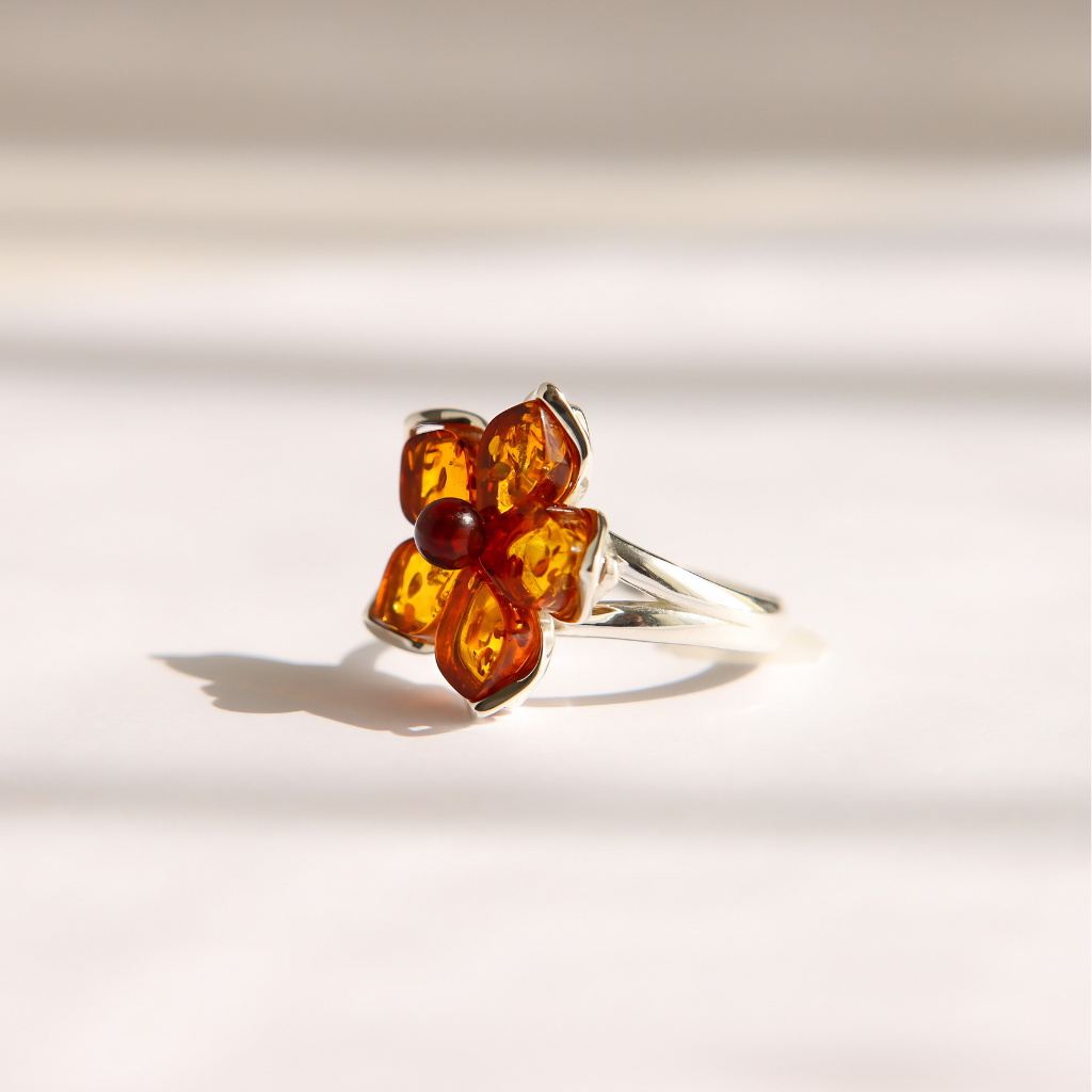 A silver amber flower ring featuring a flower-shaped setting with amber-colored stones and a small dark red center stone, photographed on a light beige surface with soft shadows.