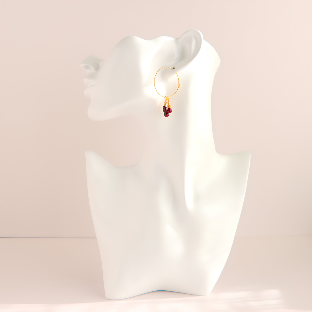 A single gold hoop earring with a dangling cluster of small red beads, displayed on a white female bust against a soft beige background.
