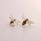 Silver bee-shaped amber earrings with orange and black details on a light pink background