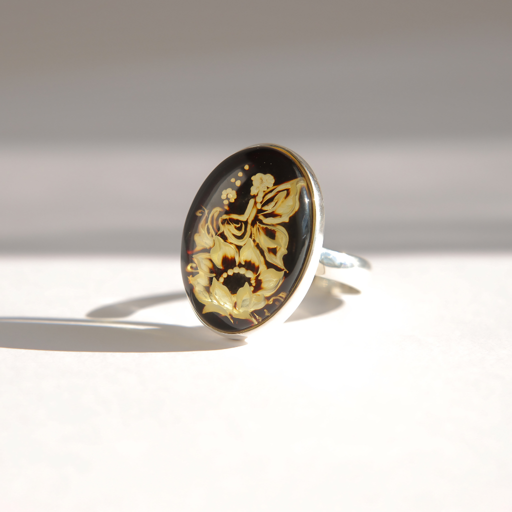 An oval amber cameo ring featuring a black cabochon stone with a detailed carving of a fairy and floral design, set in a silver band on a light gray background.