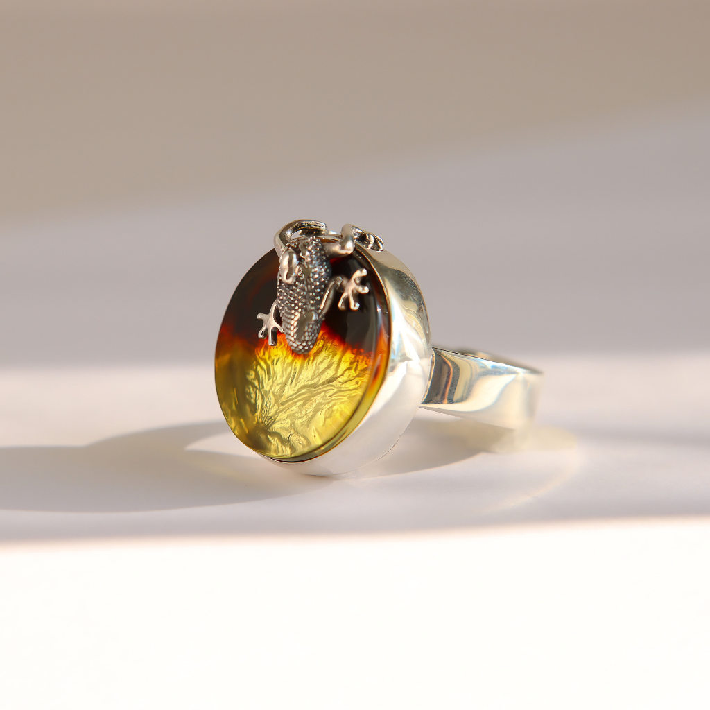 A silver amber ring featuring a large oval cabochon with a natural amber and dark brown gradient, detailed with a golden-yellow floral pattern near the bottom and a small silver frog figure perched on top, set against a clean white background with soft shadows.