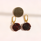 A pair of amber rose earrings with dark reddish-brown carved petals and gold leverback hooks, shown next to a US quarter coin for scale on a light beige background.