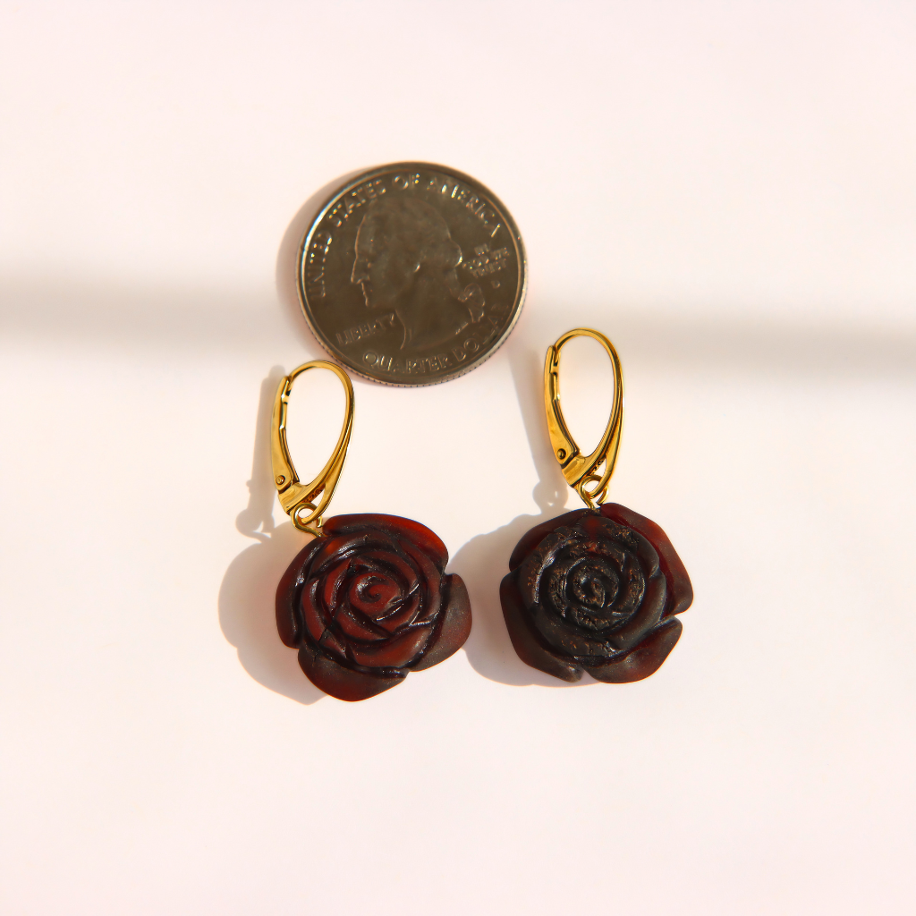 A pair of amber rose earrings with dark reddish-brown carved petals and gold leverback hooks, shown next to a US quarter coin for scale on a light beige background.