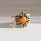 A silver and gold-toned metal amber cuff bracelet with an ornate central cross-like motif and a round orange cabochon stone, photographed on a light gray background.