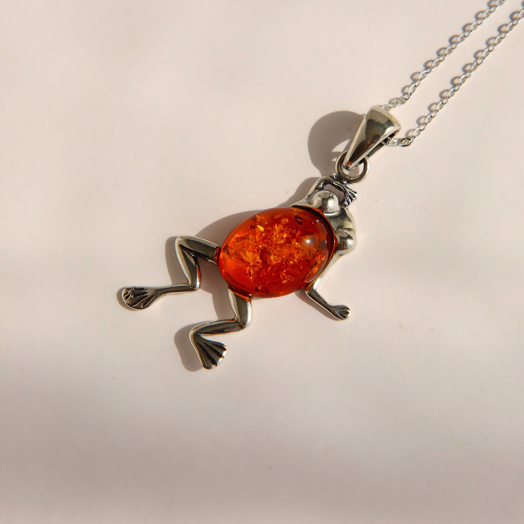 A silver amber frog pendant necklace with a vibrant orange amber-filled body, shown on a plain light beige background.