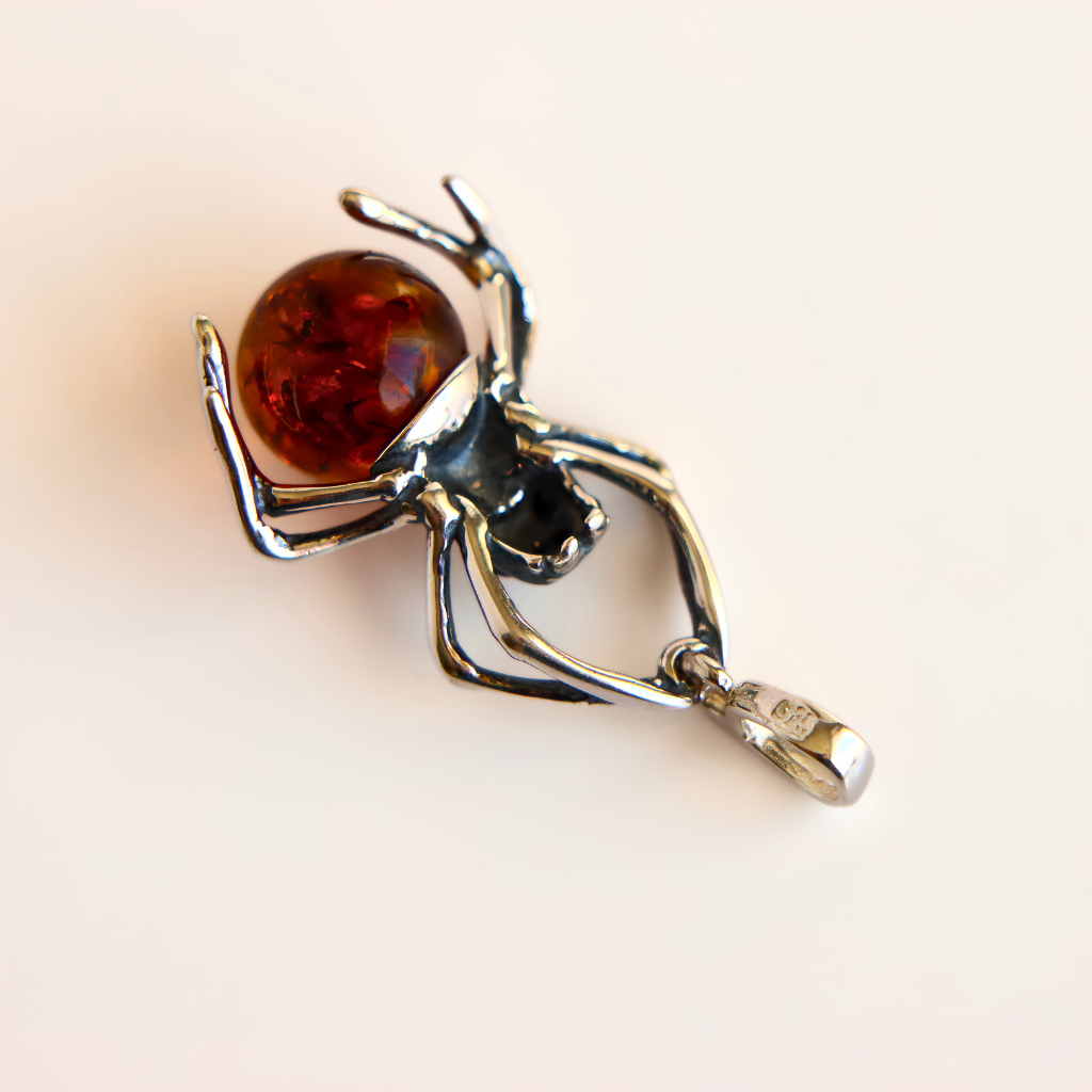 A amber spider pendant charm made of silver-toned metal with a faceted amber-brown orb on its abdomen, photographed on a plain light beige background.