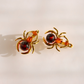 A pair of gold spider-shaped earrings with dark red and orange gemstone accents, displayed on a clean white background.