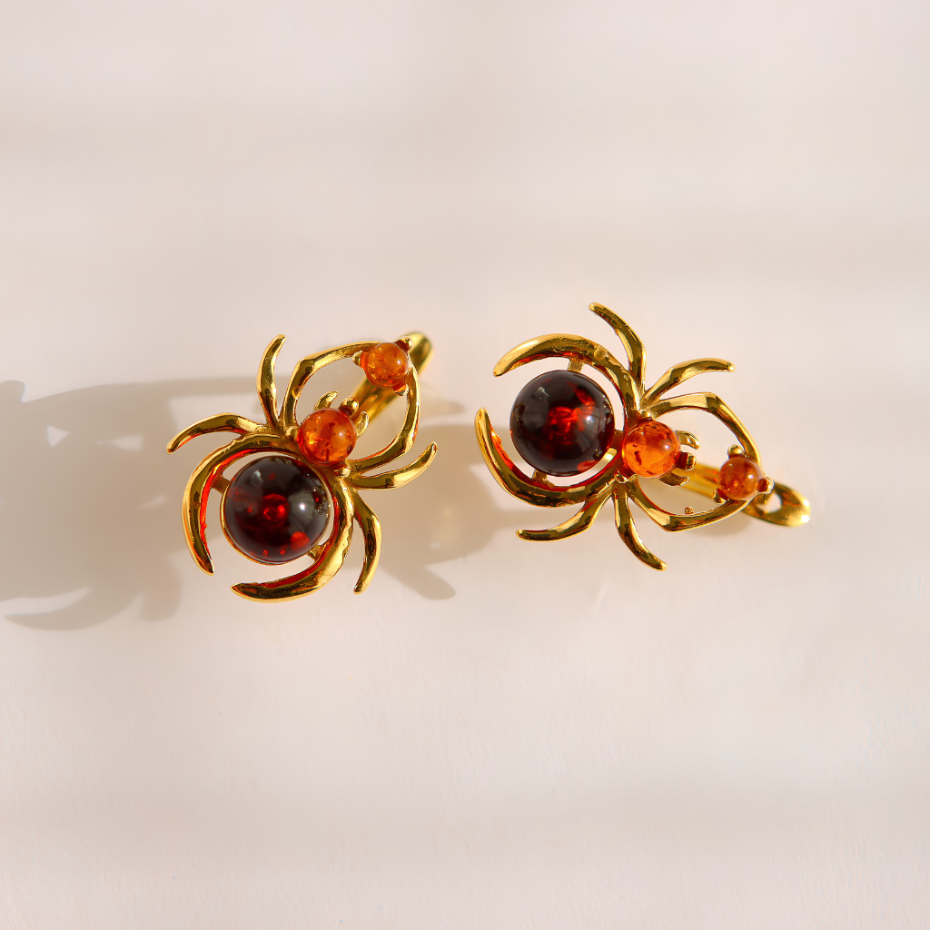 A pair of gold spider-shaped earrings with dark red and orange gemstone accents, displayed on a clean white background.