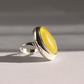 A sterling silver amber poison ring with a large, oval, yellow cabochon gemstone, photographed on a light gray surface with soft shadows.