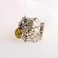 A silver spider amber bracelet with an intricate web design and a round yellow-green stone, photographed on a clean white background.