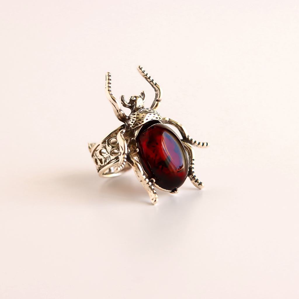 A silver scarab ring with a large, polished dark red oval gemstone on a light cream background.