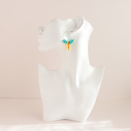 A pair of small turquoise hoop earrings with turquoise blue and orange dangling beads, displayed on a white mannequin head against a soft beige background.