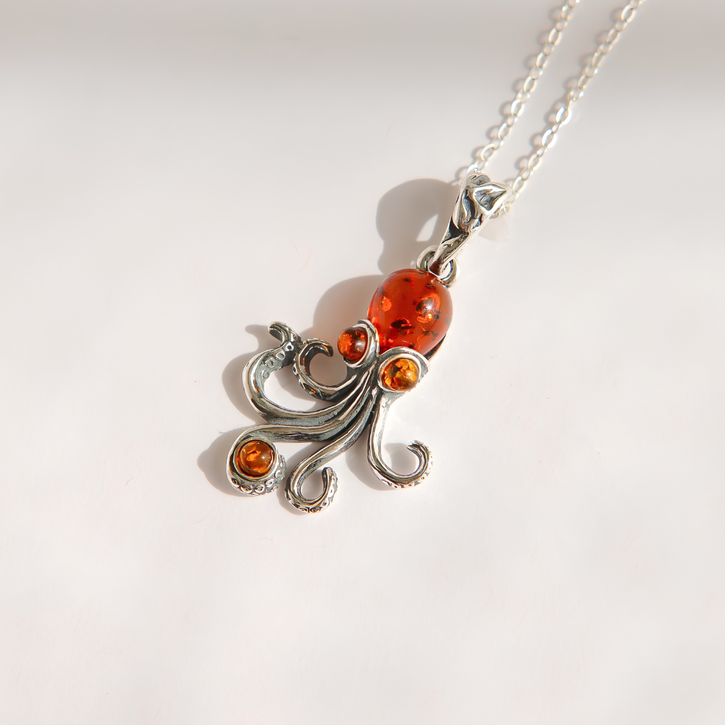 A silver amber pendant necklace featuring a stylized octopus design set with amber-colored stones, including a large round amber cabachon and two smaller accent stones, on a silver chain against a plain white background.