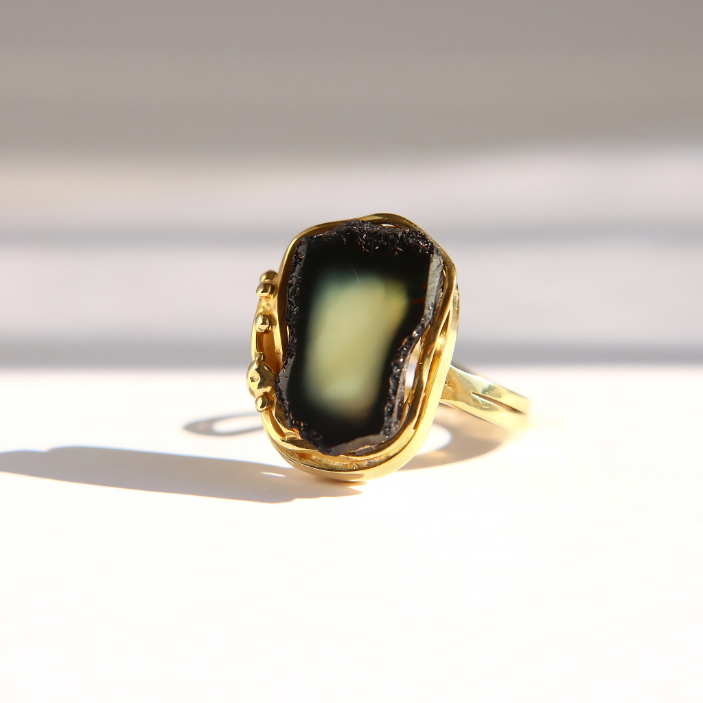 A gold amber ring featuring a large, irregular-shaped black amber stone with a pale greenish-yellow center, photographed on a white surface with a soft shadow.