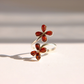 A silver Baltic amber flower ring featuring two flower charms, each with four amber-red teardrop stones, photographed on a warm off-white surface with soft shadows.