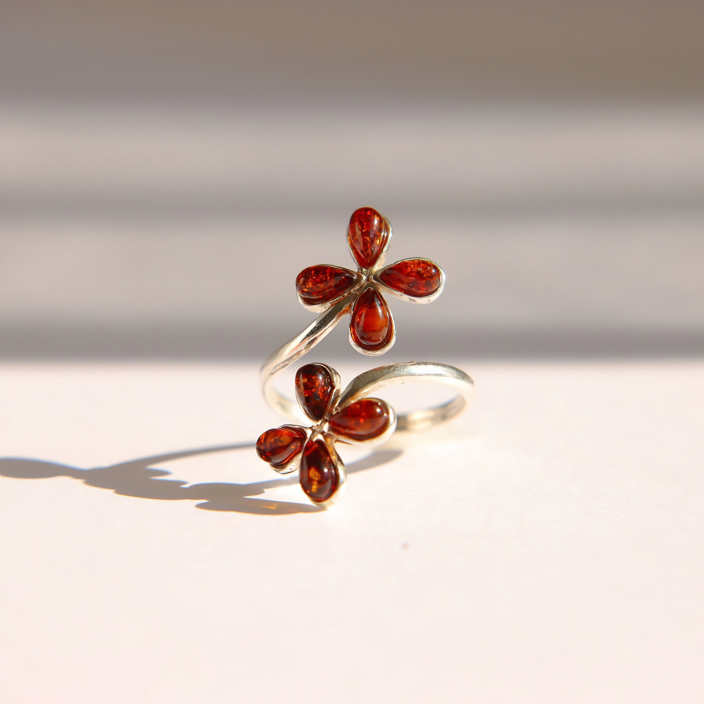 A silver Baltic amber flower ring featuring two flower charms, each with four amber-red teardrop stones, photographed on a warm off-white surface with soft shadows.