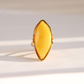 A gold butterscotch amber ring featuring a large, marquise-shaped yellow stone, photographed on a light beige surface with soft shadows.