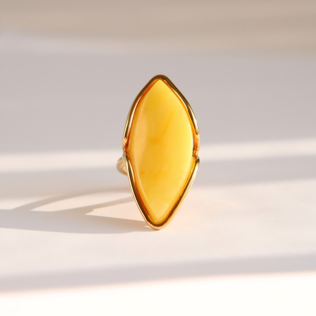 A gold butterscotch amber ring featuring a large, marquise-shaped yellow stone, photographed on a light beige surface with soft shadows.