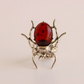 A scarab ring featuring a polished oval red gemstone on a textured silver-toned metal body with detailed legs and antennae, set against a soft beige background.