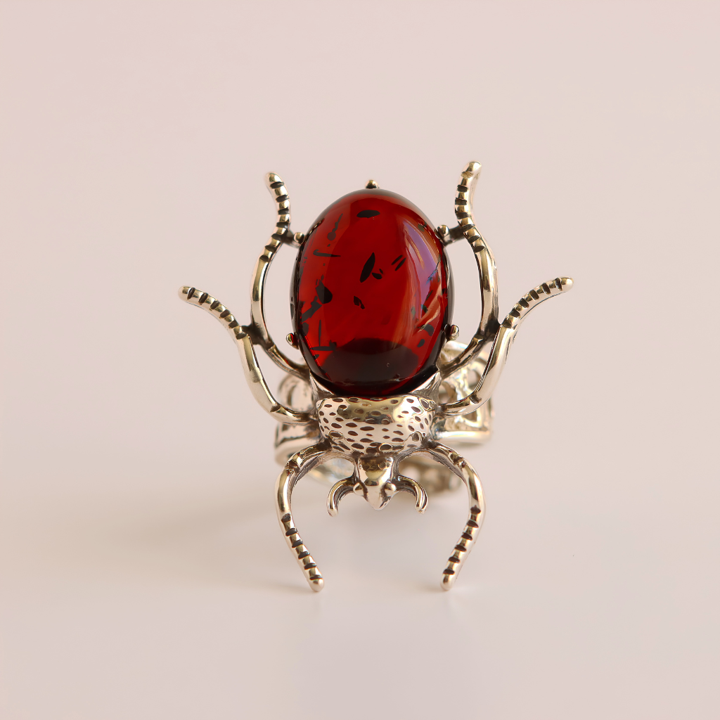 A scarab ring featuring a polished oval red gemstone on a textured silver-toned metal body with detailed legs and antennae, set against a soft beige background.