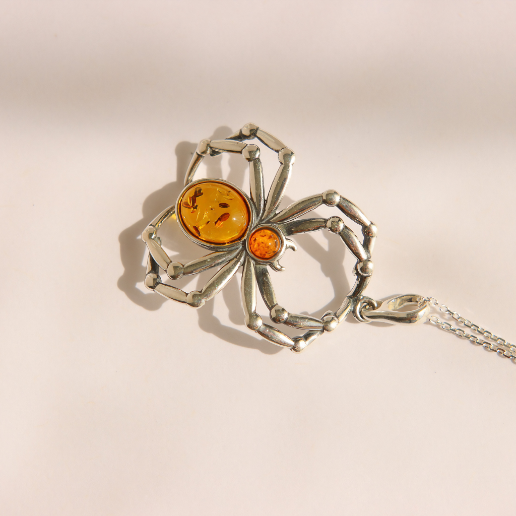 A silver amber spider pendant necklace with two amber-colored gemstones, displayed on a light beige background with a soft shadow.