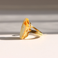 A gold white amber ring with a large, irregularly shaped cream-colored stone featuring orange-brown marbling, photographed on a white surface with soft diagonal shadows.
