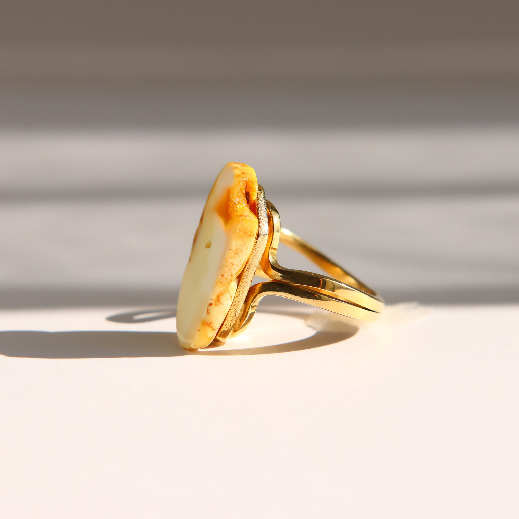 A gold white amber ring with a large, irregularly shaped cream-colored stone featuring orange-brown marbling, photographed on a white surface with soft diagonal shadows.