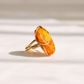 A gold amber ring featuring a large, irregularly shaped amber-orange stone, photographed on a clean white surface with soft, diagonal shadows.