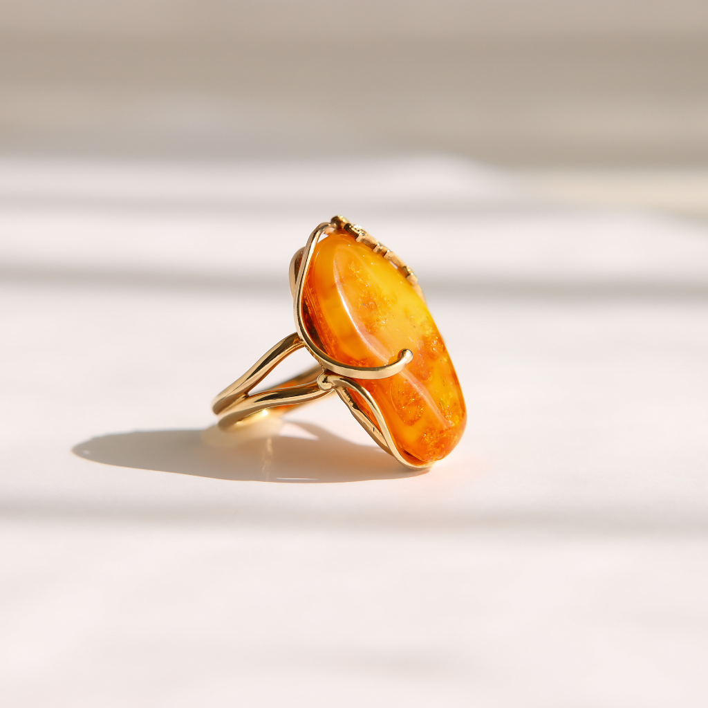 A gold amber ring featuring a large, irregularly shaped amber-orange stone, photographed on a clean white surface with soft, diagonal shadows.