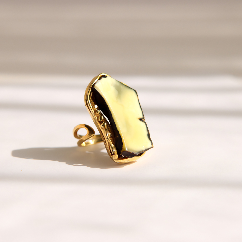 A gold amber statement ring with an irregular, polished stone face, photographed on a light beige background with soft diagonal shadows.