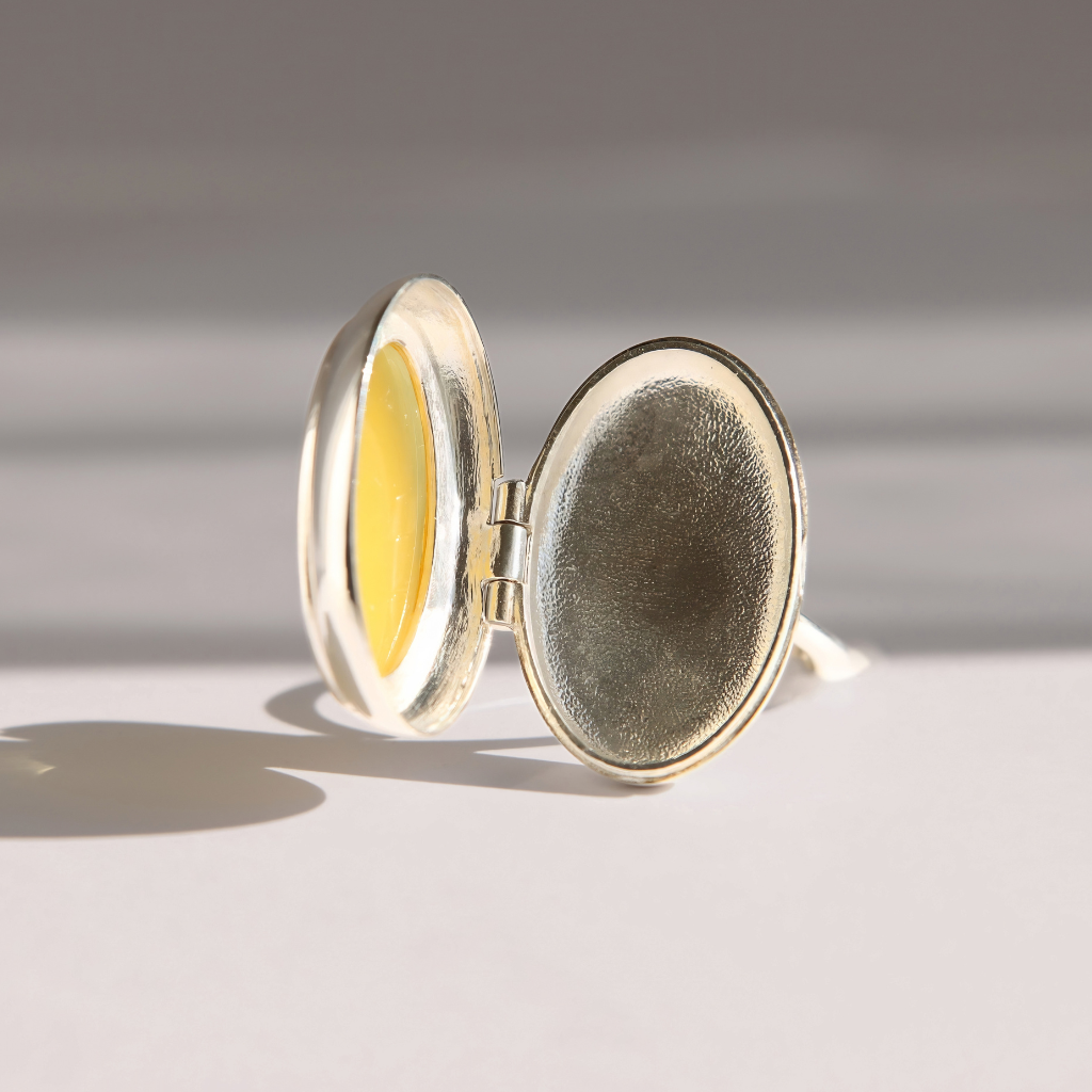 A silver oval amber poison ring shown open, with a yellow interior lining and a textured silver exterior, photographed on a clean light gray surface with soft shadows.