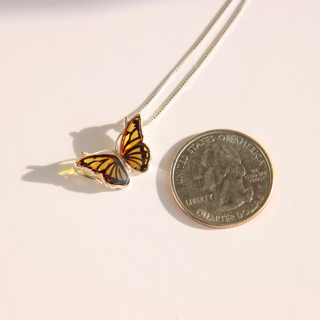 A silver amber butterfly pendant necklace with orange and black patterned wings, shown next to a US quarter coin for scale on a white background.