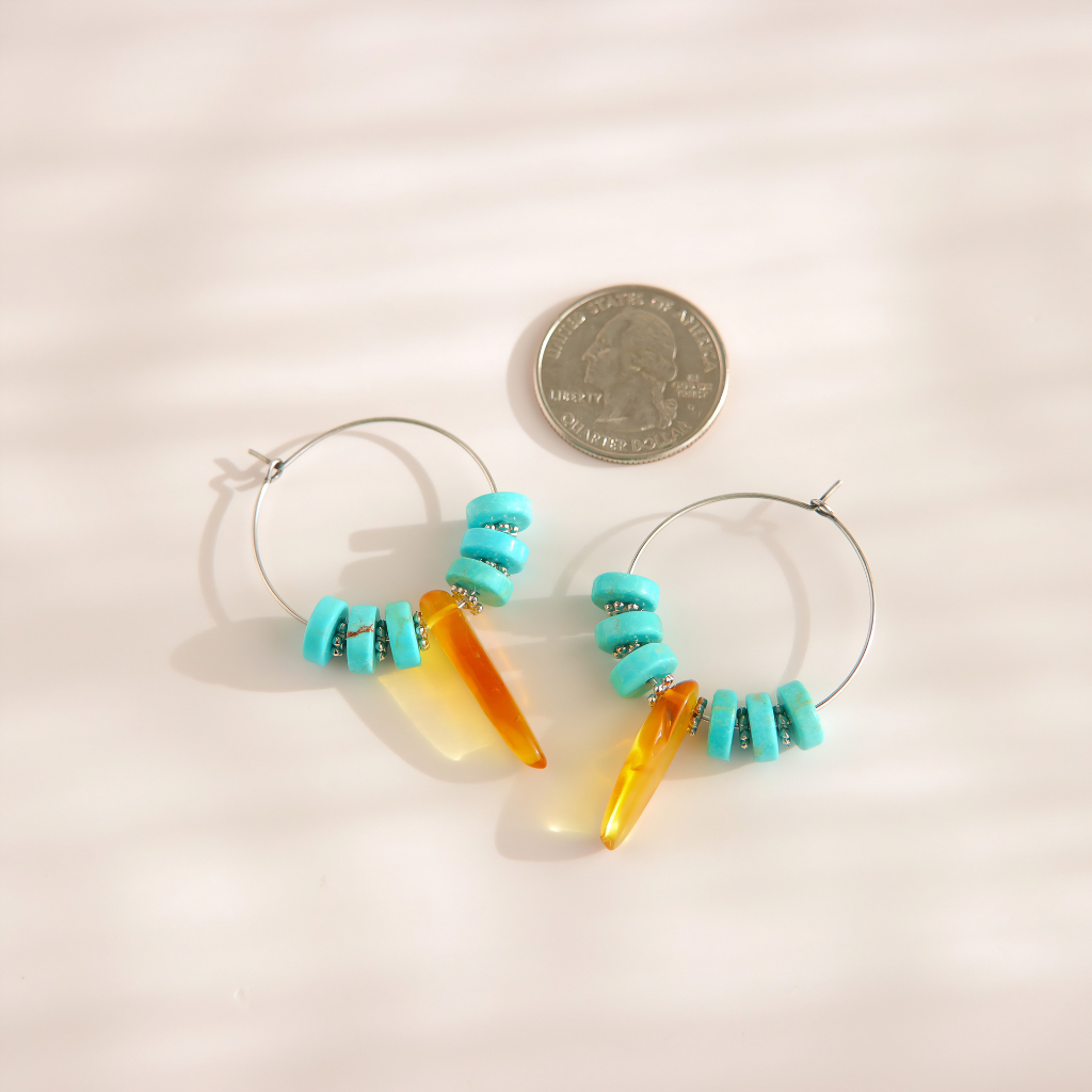A pair of silver and turquoise hoop earrings with turquoise beads and amber-colored dangling charms, shown next to a US quarter coin for scale on a light beige background with soft shadows.